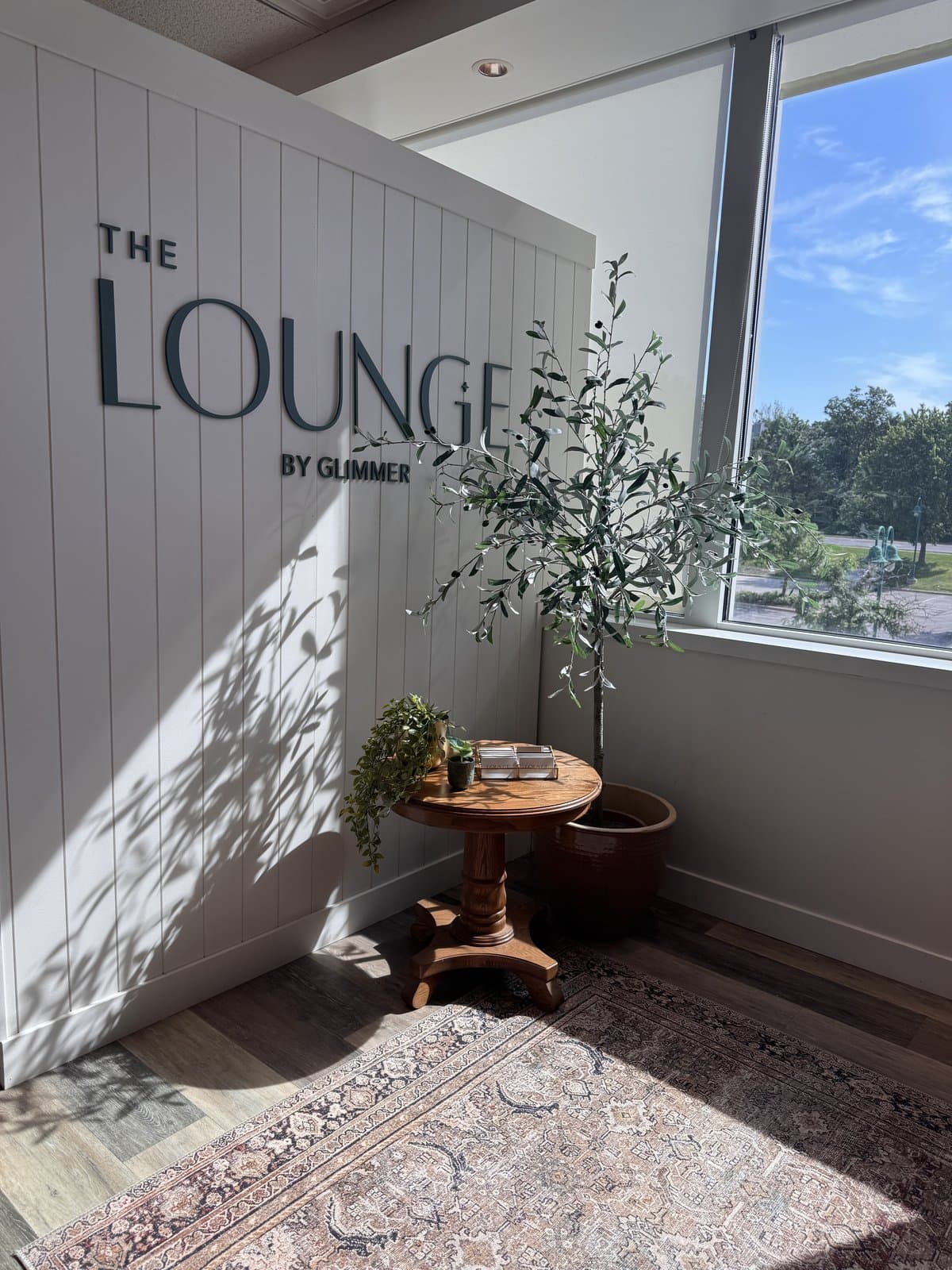The Lounge by Glimmer wordmark wall with an olive tree by the window